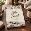 A hardcover book or journal titled "John Smith" with a beige cover and torn newspaper-style borders, placed on a wooden table in a cozy indoor setting with a coffee cup, glasses, and plants nearby.