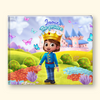 A personalized children's picture book cover featuring a cartoon prince in a blue uniform and gold crown standing in a colorful fantasy landscape with a castle, flowers, butterflies, and the text 'Jamie the prince'. The book is photographed upright against a light cream background.